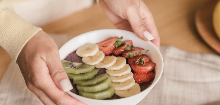 Delicious smoothie bowl topped with fresh fruits like kiwi, banana, and strawberries, held by manicured hands.