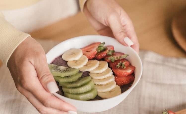Delicious smoothie bowl topped with fresh fruits like kiwi, banana, and strawberries, held by manicured hands.